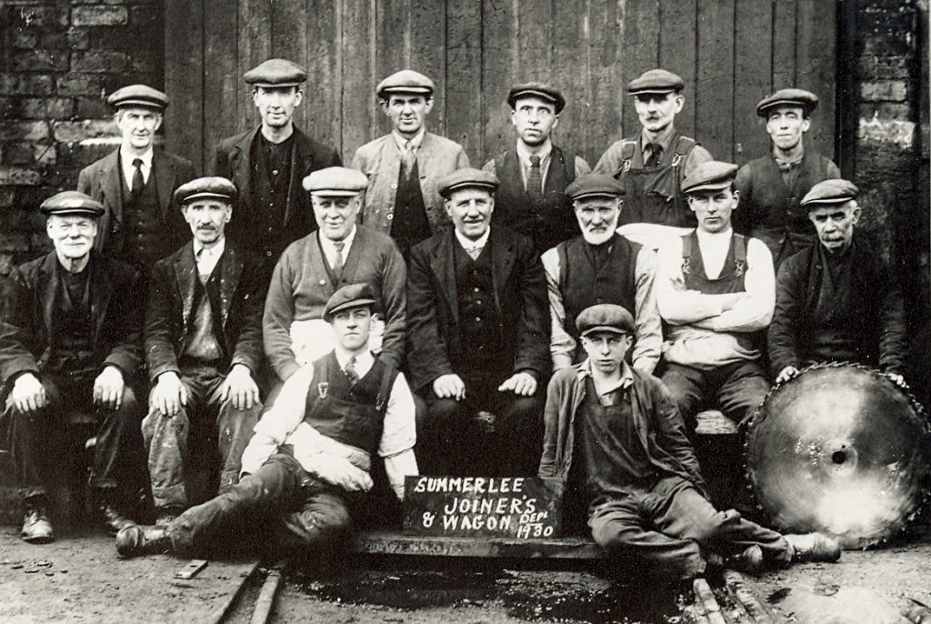 Black & white photograph of 15 men in overalls and caps posed around a sign painted "Summerlee Joiners & Wagon Dept 1930"