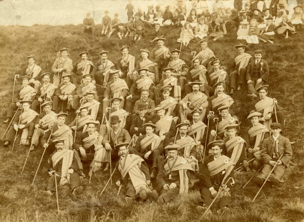 Sepia toned photograph of a large group of men sitting on a hillside dressed in bonnets and plaids and holding shepherds crooks. A smaller group of women and children can be seen above them.