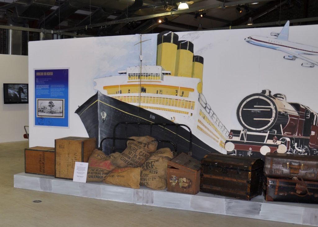 An exhibition set display of trunks, suitcases, crates and sugar sacks against the backdrop of a mural featuring a transatlantic liner, steam train and aeroplane