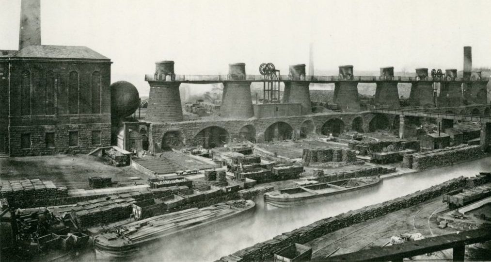 A LANDSCAPE FORMAT BLACK AND WHITE PHOTO LOOKING ACROSS A CANAL TO A ROW OF IRON FURNACES WITH A LARGE EGINE HOUSE AND CHIMNEY ON THE LEFT. TWO BARGES ARE TIED UP ON THE CANAL AND A TRAIN OF RAILWAY WAGONS SITS NEARBY. PHOTO TAKEN IN THE 1870s.