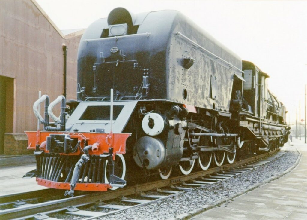 View of a large, industrial steam locomotive with one of its two tenders in the foreground. The loco is painted black but has a red cow-catcher at the end. There is a large warehouse-like building in the background.